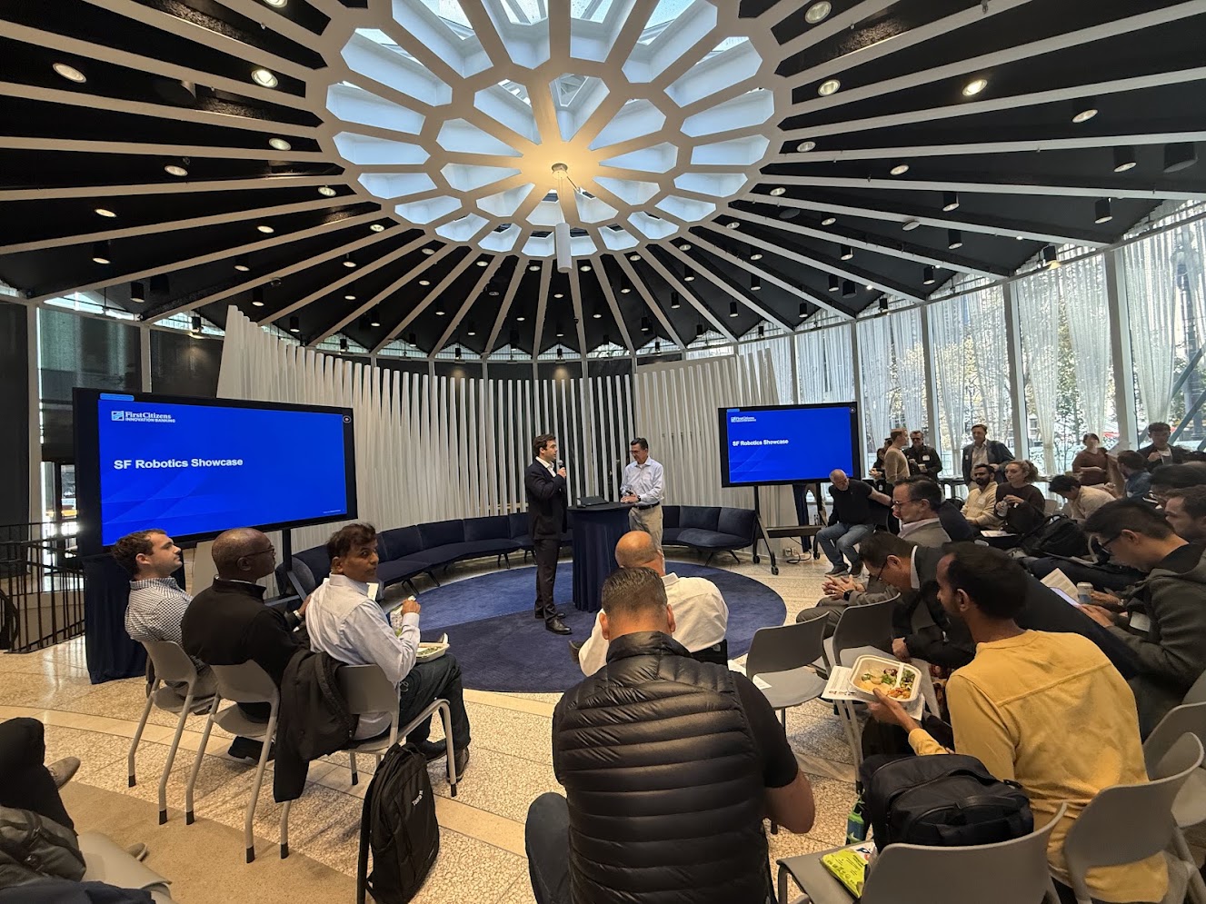 The SF Robotics Showcase — two hosts speaking at the front of a round room with a geometric skylight, blue screens reading SF Robotics Showcase, and an audience seated at tables